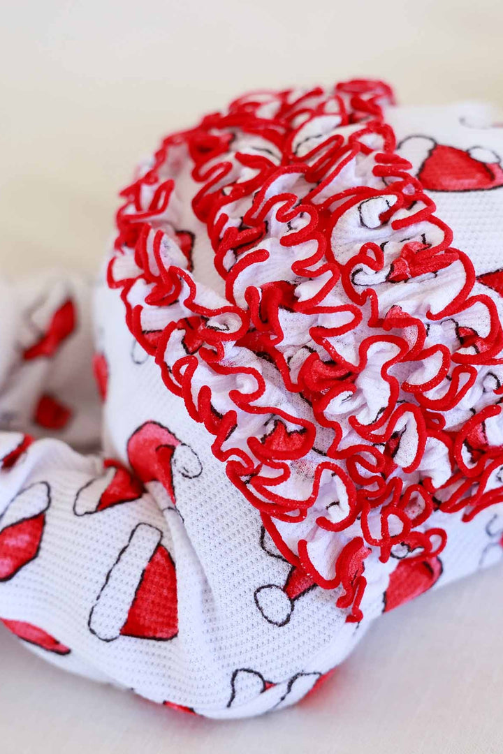 Baby wearing a Caden Lane Santa hat print ruffle footie while lying on a soft white blanket