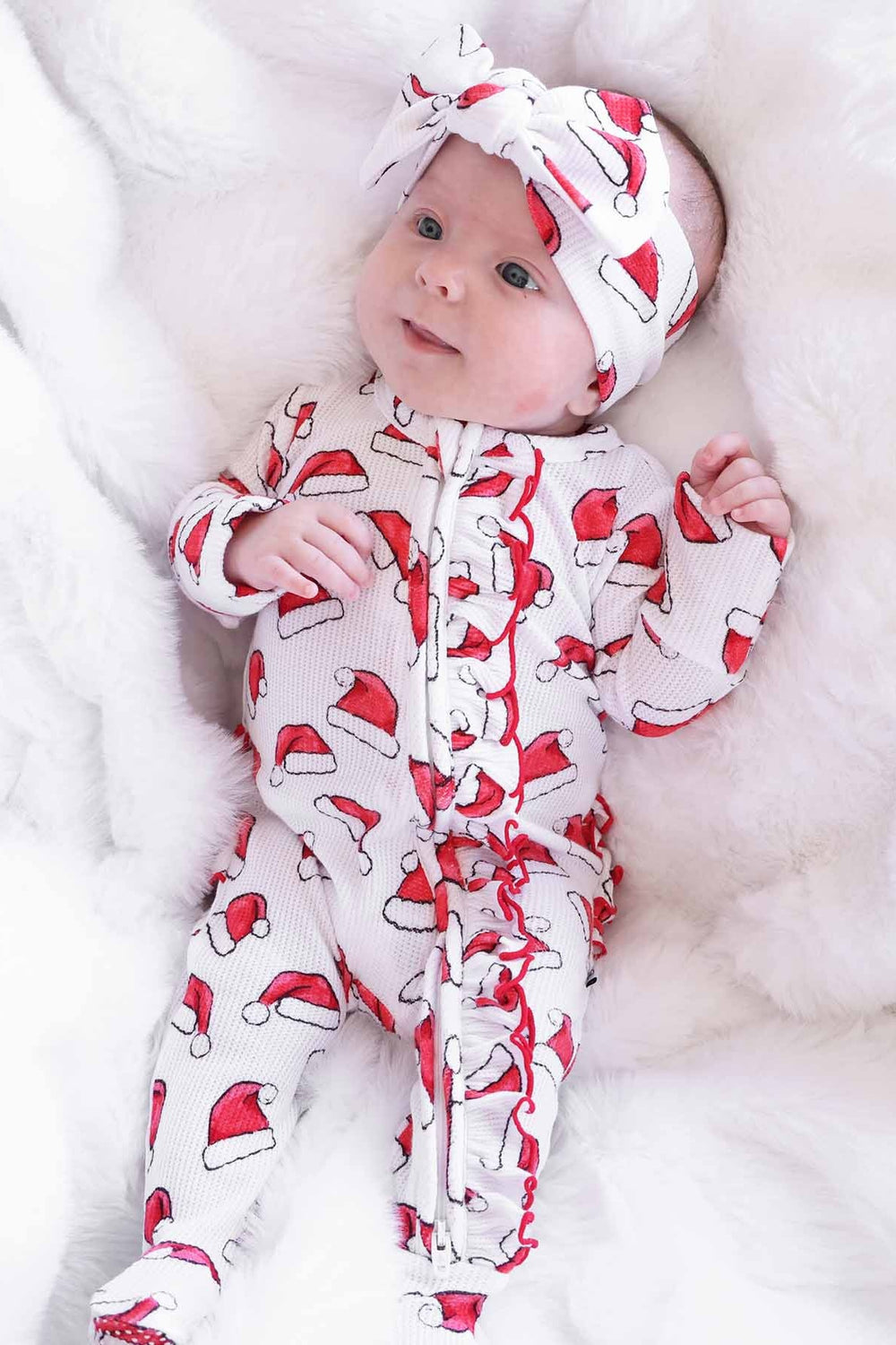 Baby wearing a Caden Lane Santa hat print ruffle footie with a matching knotted headband while lying on a soft white blanket