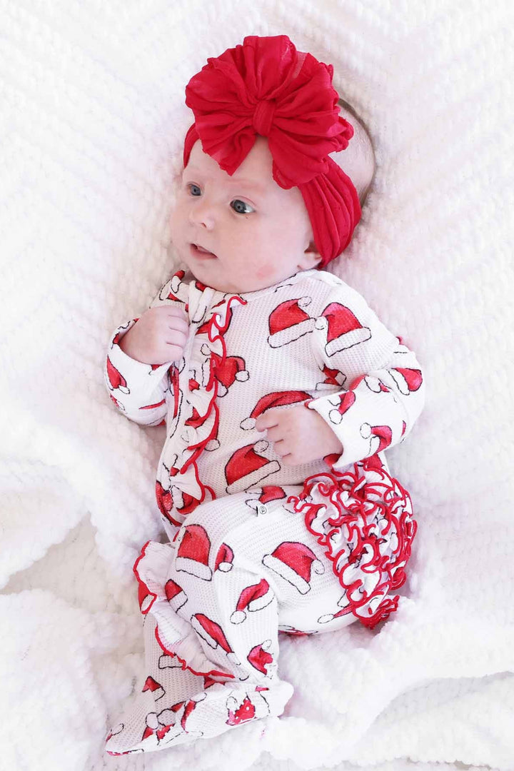 Baby wearing a Caden Lane Santa hat print ruffle footie with a large red bow headband while lying on a soft white blanket