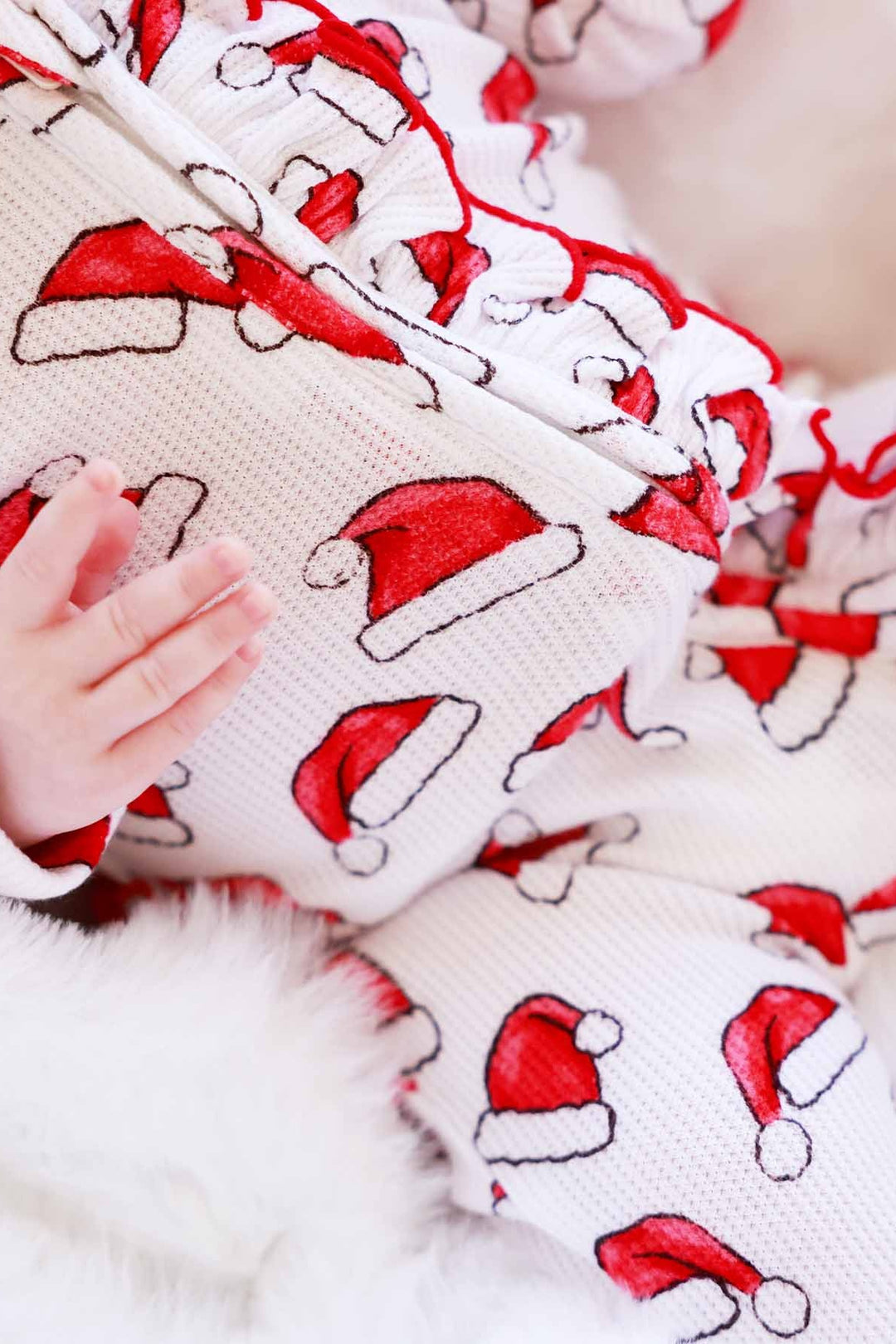 Baby wearing a Caden Lane Santa hat print ruffle footie while lying on a soft white blanket