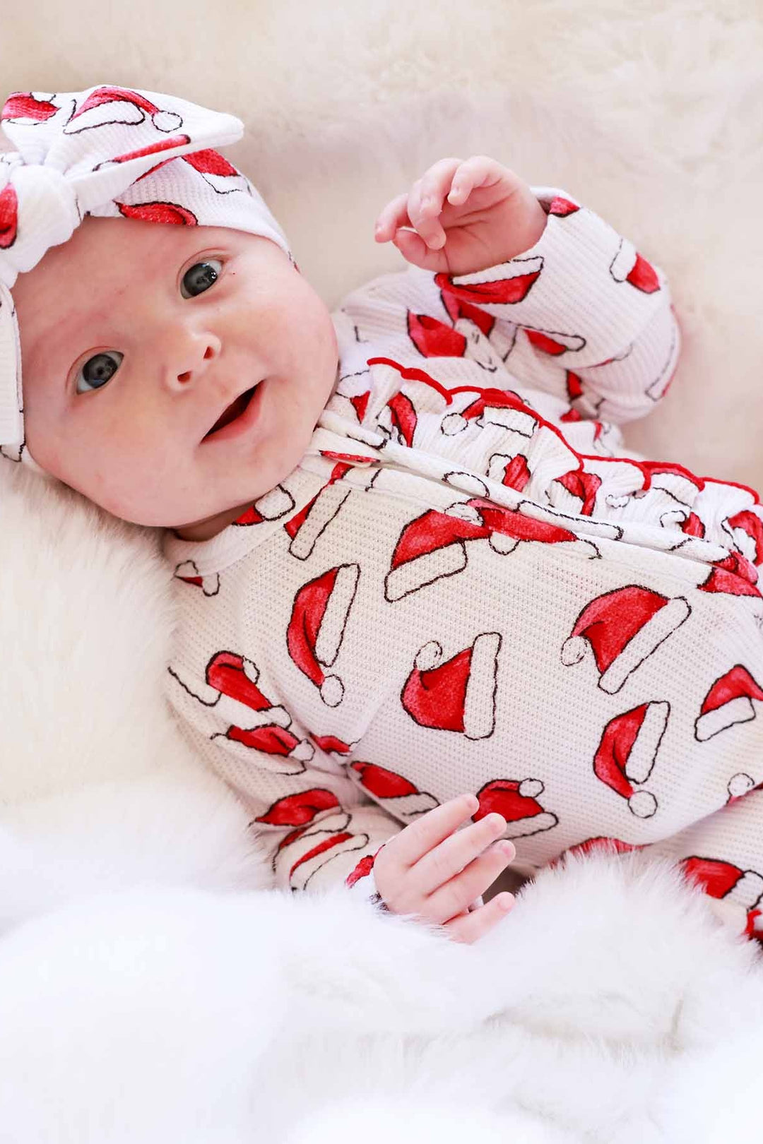 Baby wearing a Caden Lane Santa hat print ruffle footie with a matching knotted headband while lying on a soft white blanket