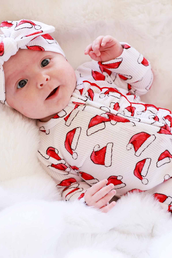 Baby wearing a Caden Lane Santa hat print ruffle footie with a matching knotted headband while lying on a soft white blanket
