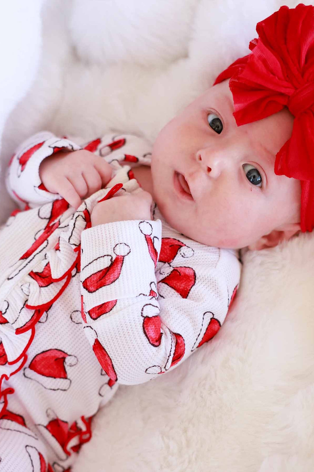 Baby wearing a Caden Lane Santa hat print ruffle footie with a large red bow headband while lying on a soft white blanket