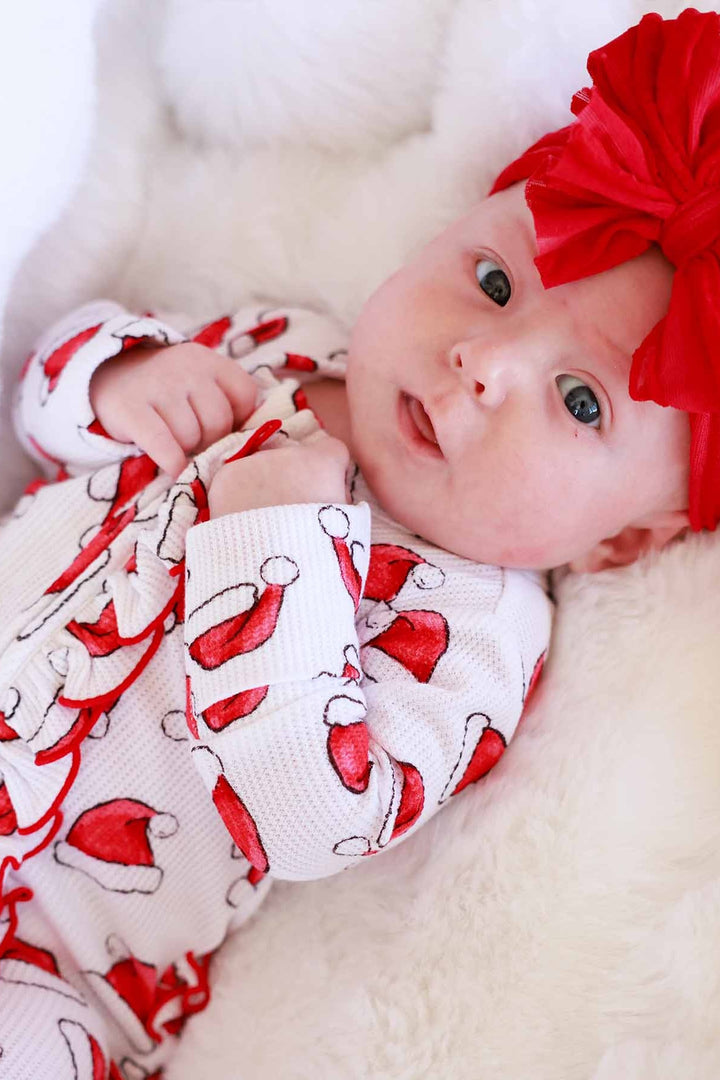 Baby wearing a Caden Lane Santa hat print ruffle footie with a large red bow headband while lying on a soft white blanket