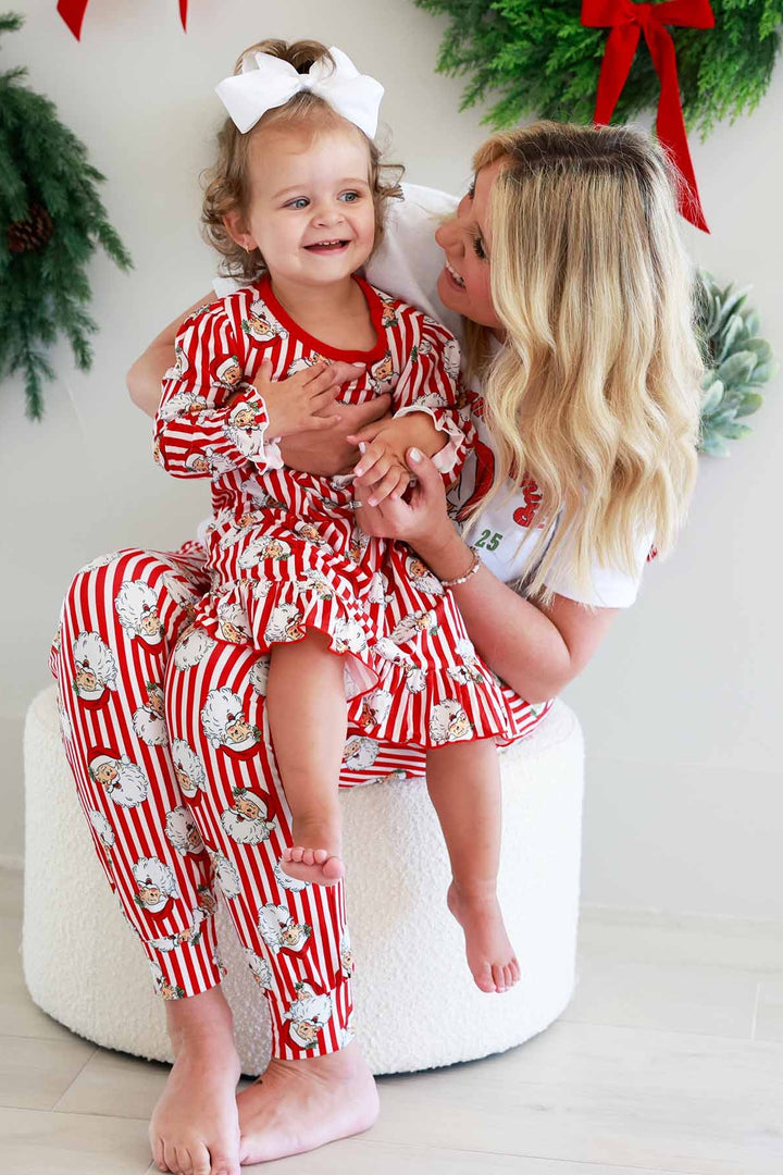 Mother and daughter in matching red-and-white Caden Lane striped Christmas pajamas with a vintage Santa print. The mom holds her smiling toddler while sitting on a white ottoman, surrounded by festive holiday wreaths with red bows, showcasing coordinated family Christmas sleepwear.