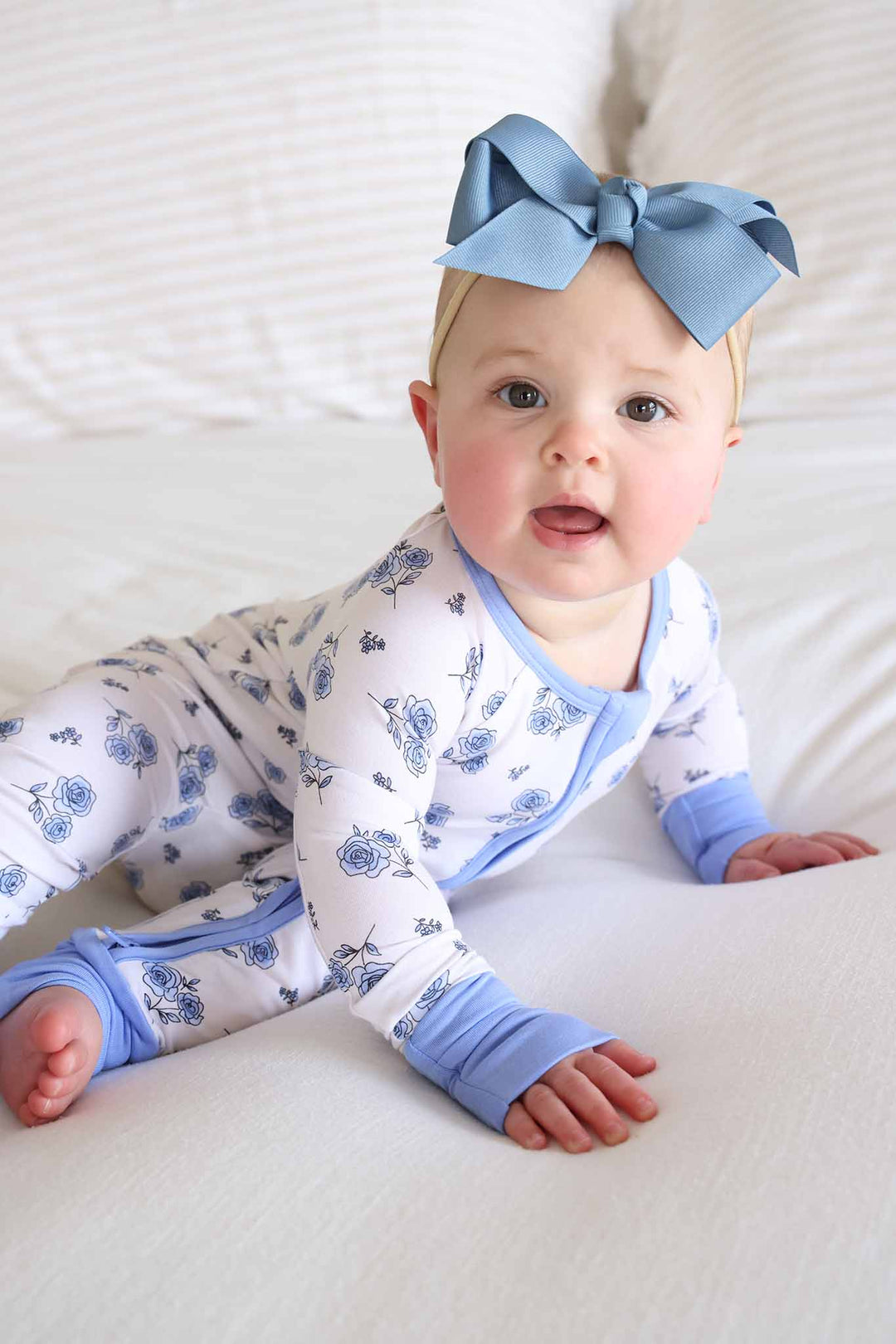 baby girl leaning over to the size wearing a blue rose convertible zip romper pajama with a blue ribbon bow headband 