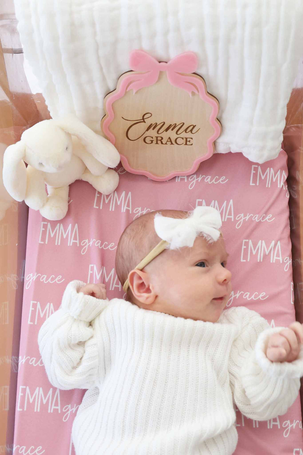 baby girl laying in bassinet wearing a white sweater set with a matching white bow on a pink personalized swaddle with a wooden name sign with a pink bow on it