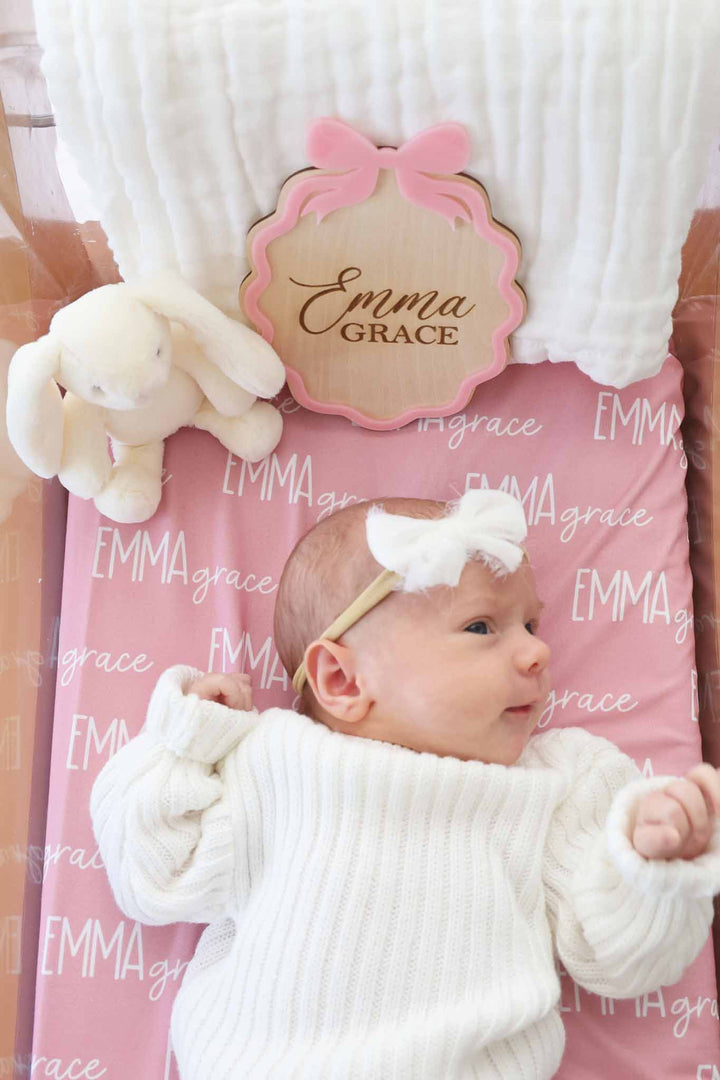 baby girl laying in bassinet wearing a white sweater set with a matching white bow on a pink personalized swaddle with a wooden name sign with a pink bow on it