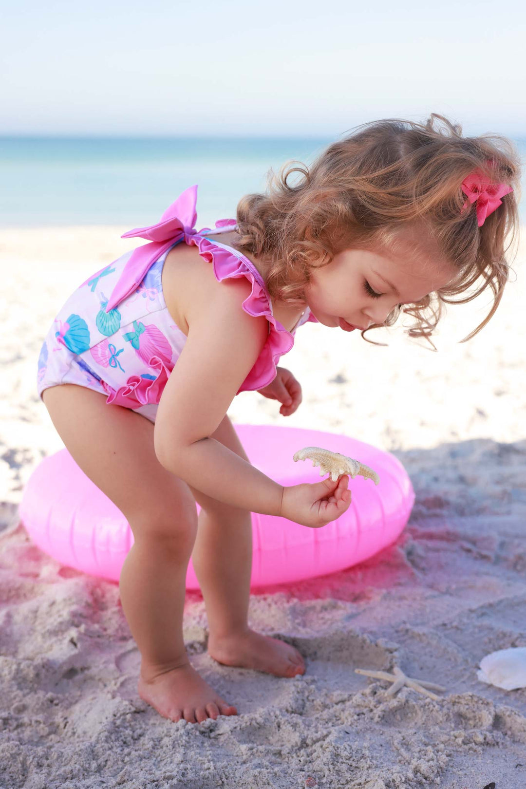 girl picking up seashell on beach in blue and pink double ruffle one piece swimsuit