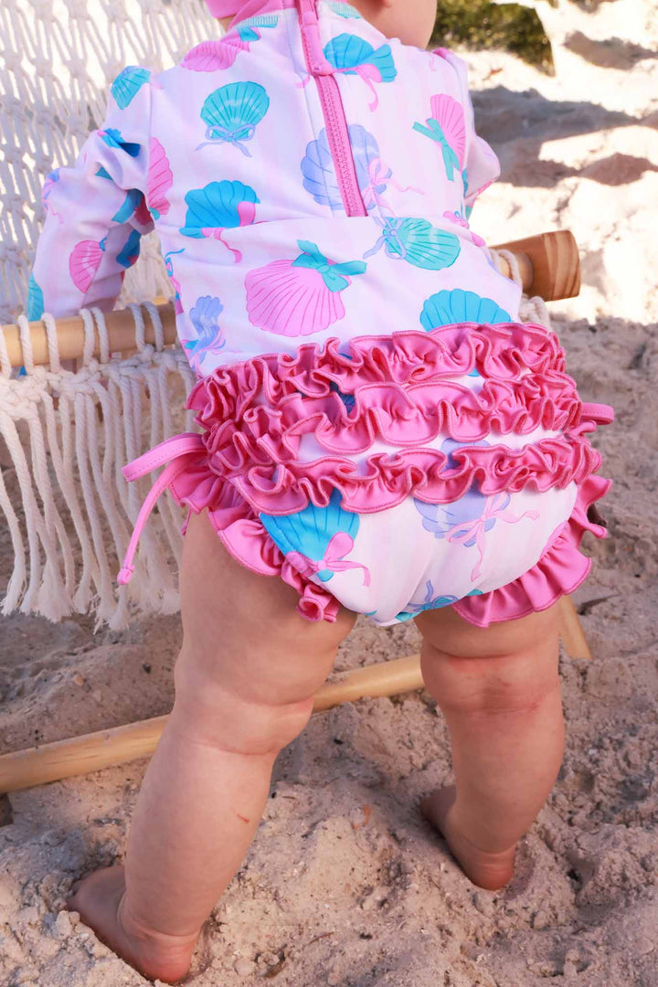 standing baby girl leaning on beach chair in seaside sweetie long sleeve rash guard with ruffle bottom 