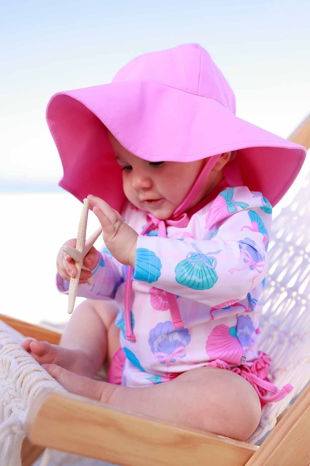 baby girl wearing a colorful seashell long sleeve rash guard swim romper with ruffles on the bottom and a matching pink swim hat