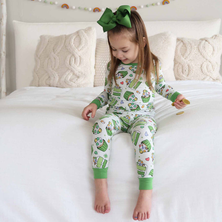 girl sitting on bed wearing a st. patrick's day pajama set with a matching green clip bow playing with gold coins 