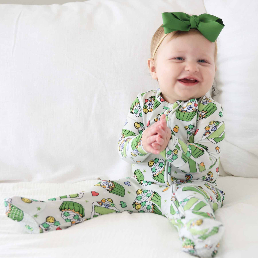 sitting baby girl wearing a green cake and cupcake footie pajama for st. patrick's day with lucky charms, clovers and pots of golf