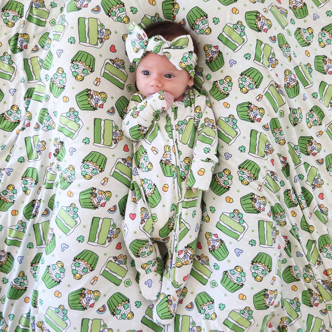 baby girls wearing a green cake and cupcake zipper footie wearing a matching headwrap and laying on a matching blanket 