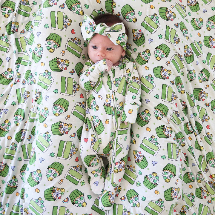 baby girls wearing a green cake and cupcake zipper footie wearing a matching headwrap and laying on a matching blanket 
