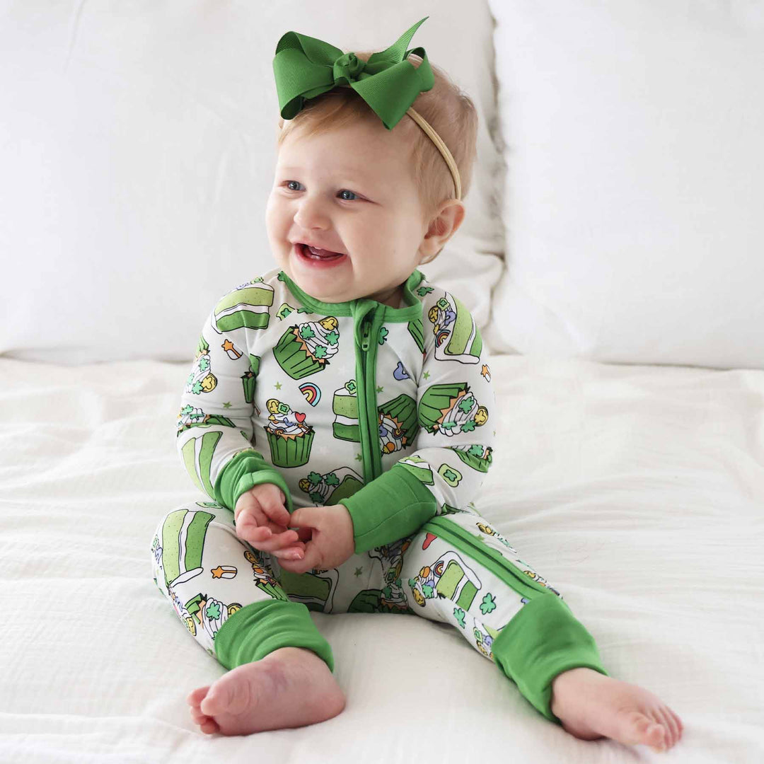 baby girl wearing a green cupcake and cake bamboo zip romper pajama with a matching green ribbon bow headband 