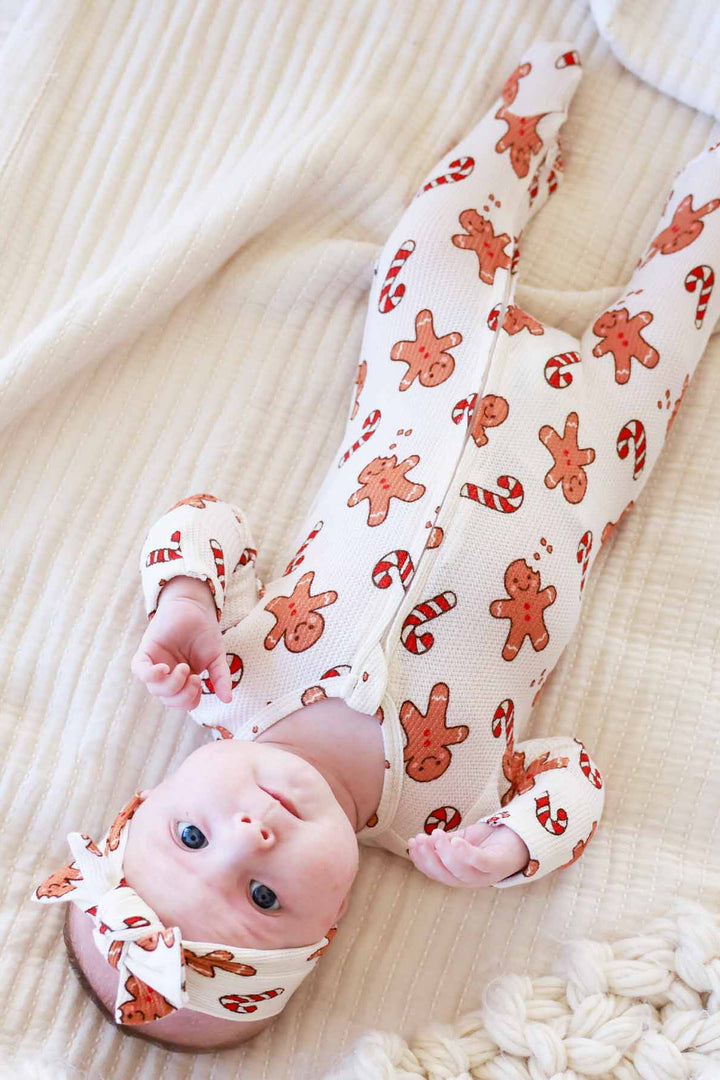 Smiling baby wearing a Caden Lane gingerbread and candy cane holiday footie pajamas on a cozy white blanket