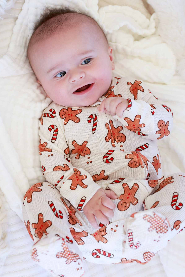 Smiling baby wearing a Caden Lane gingerbread and candy cane holiday footie pajamas on a cozy white blanket