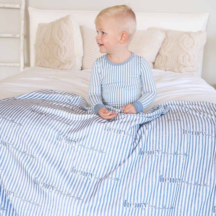 boy sitting on bed in blue striped pajama with a matching personalized blanket across his lap