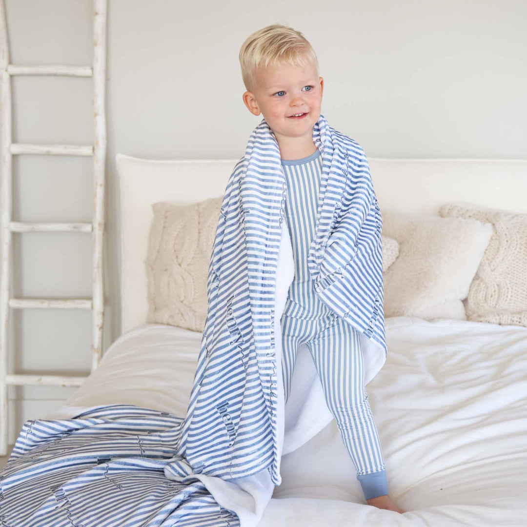 boy standing on bed wearing a blue and white stripe pajama set with a matching personalized blanket draped over his shoulders 