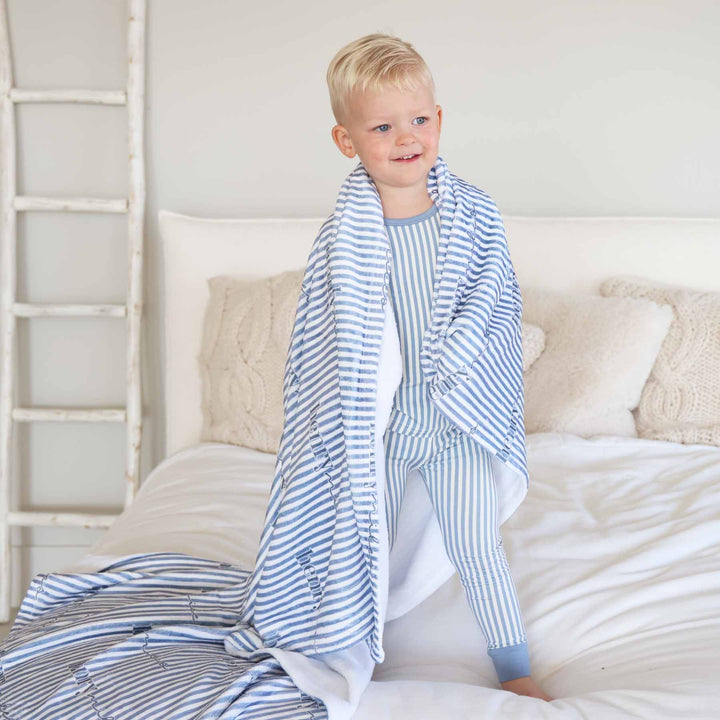 boy standing on bed wearing a blue and white stripe pajama set with a matching personalized blanket draped over his shoulders 