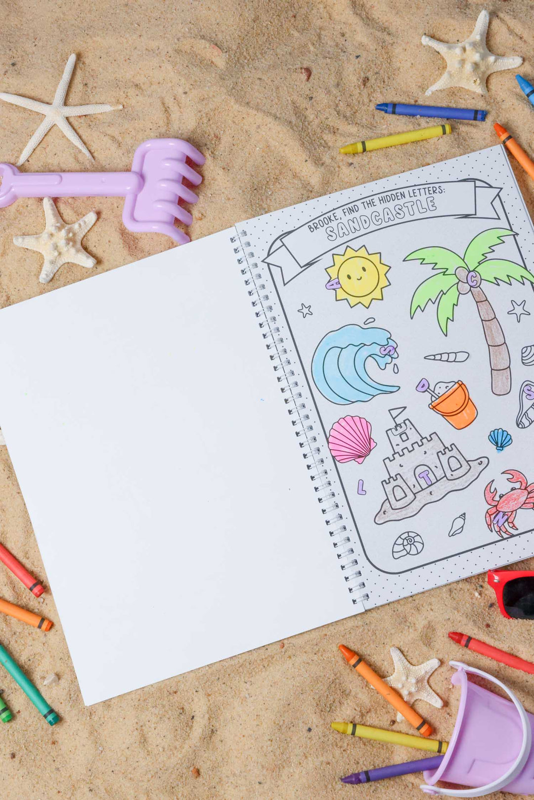 Personalized 'Brooke, find the hidden letters: SANDCASTLE' activity page from a Caden Lane coloring book, partially colored, displayed on sand with starfish and crayons.
