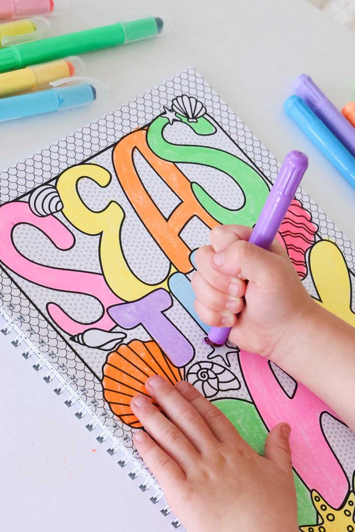 Close-up of a child's hands coloring a personalized name banner in bright colors on a mermaid-scale patterned page of a Caden Lane coloring book