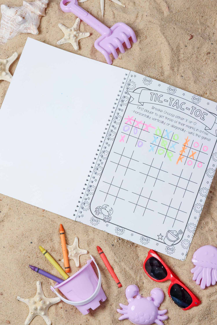 Personalized Caden Lane tic-tac-toe activity page reading 'Brooke,' partially filled in with colorful X's and O's, resting on sand with starfish, sunglasses, and beach toys.