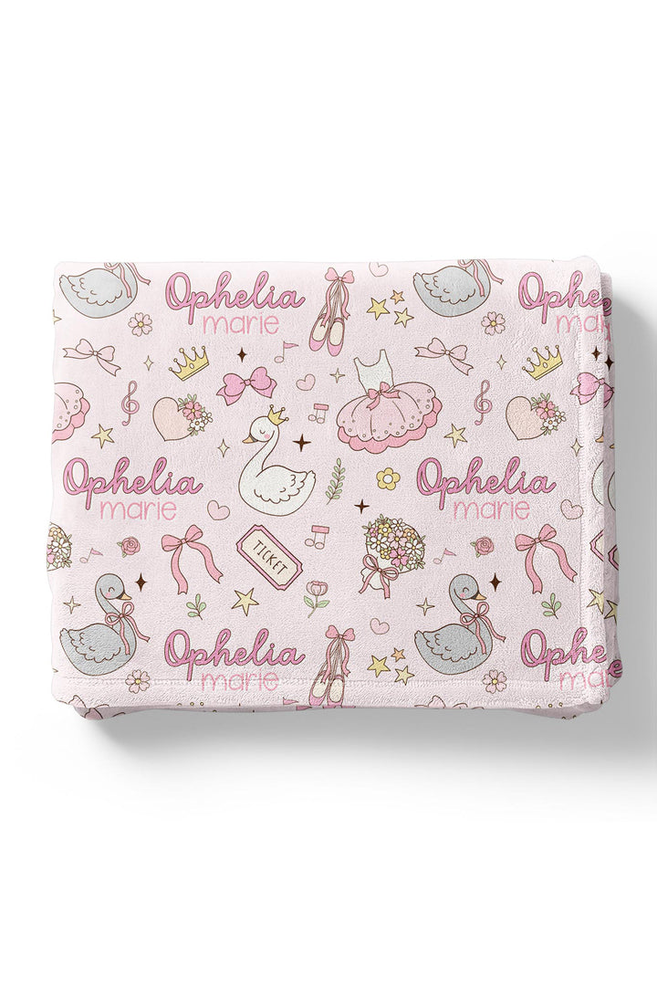 Flat lay of a folded personalized Caden Lane Swan Ballet blanket in pink, showing swans, ballet slippers, crowns, and custom name print