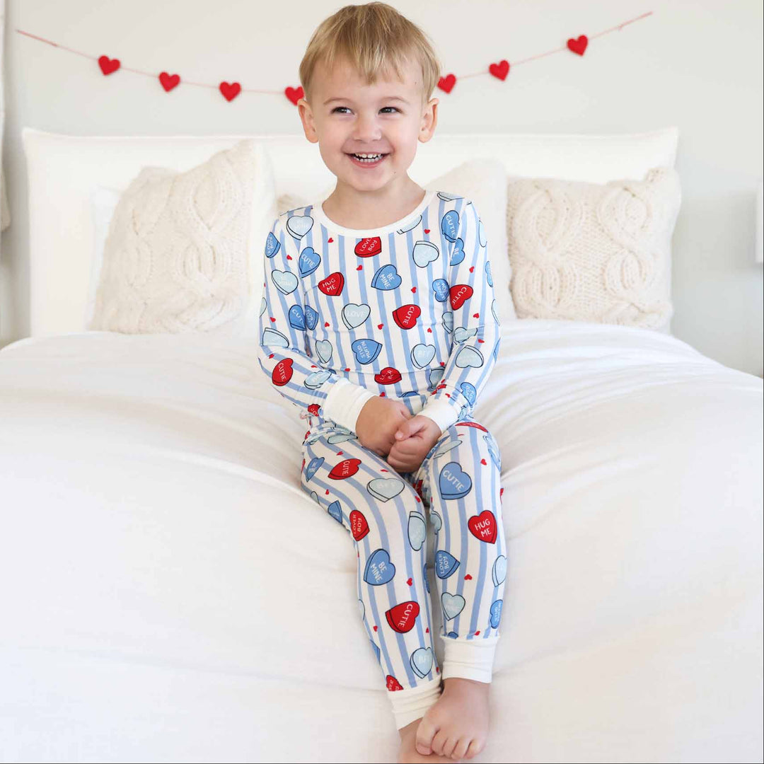 boy sitting on a bed wearing a blue striped conversation heart pajama set for kids 