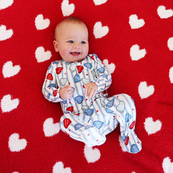 boy wearing a blue and white striped footie pajama with conversation heart candy on it laying on a red microfiber blanket with white hearts