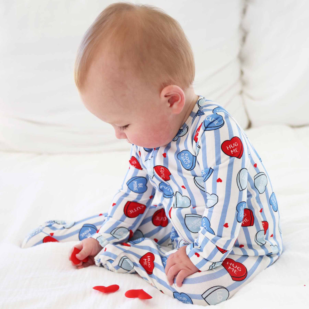 baby boy sitting on a bed playing with red heart confetti in a blue conversation heart footie pajama 