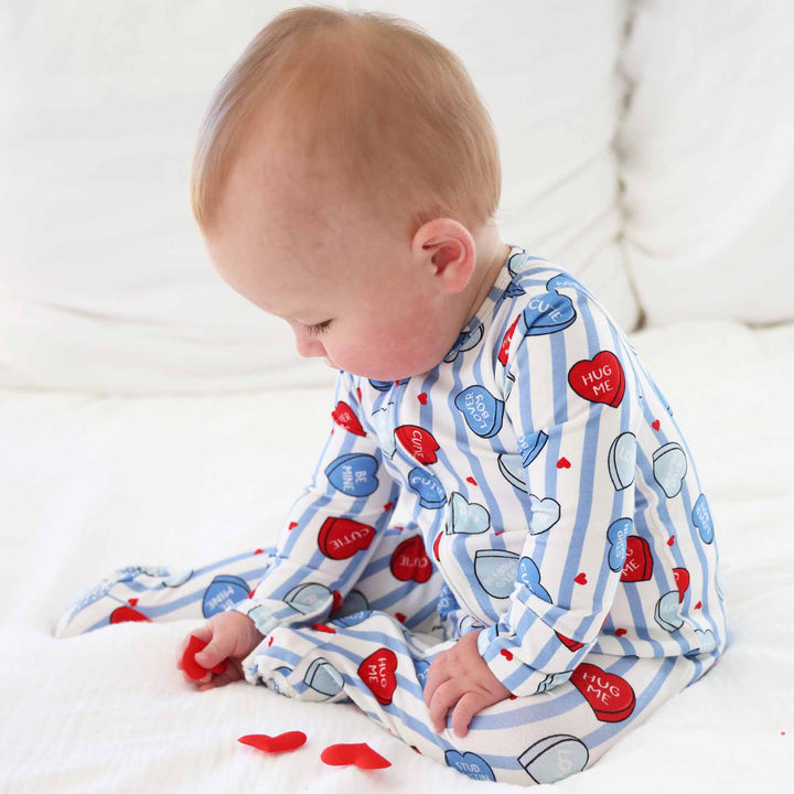 baby boy sitting on a bed playing with red heart confetti in a blue conversation heart footie pajama 