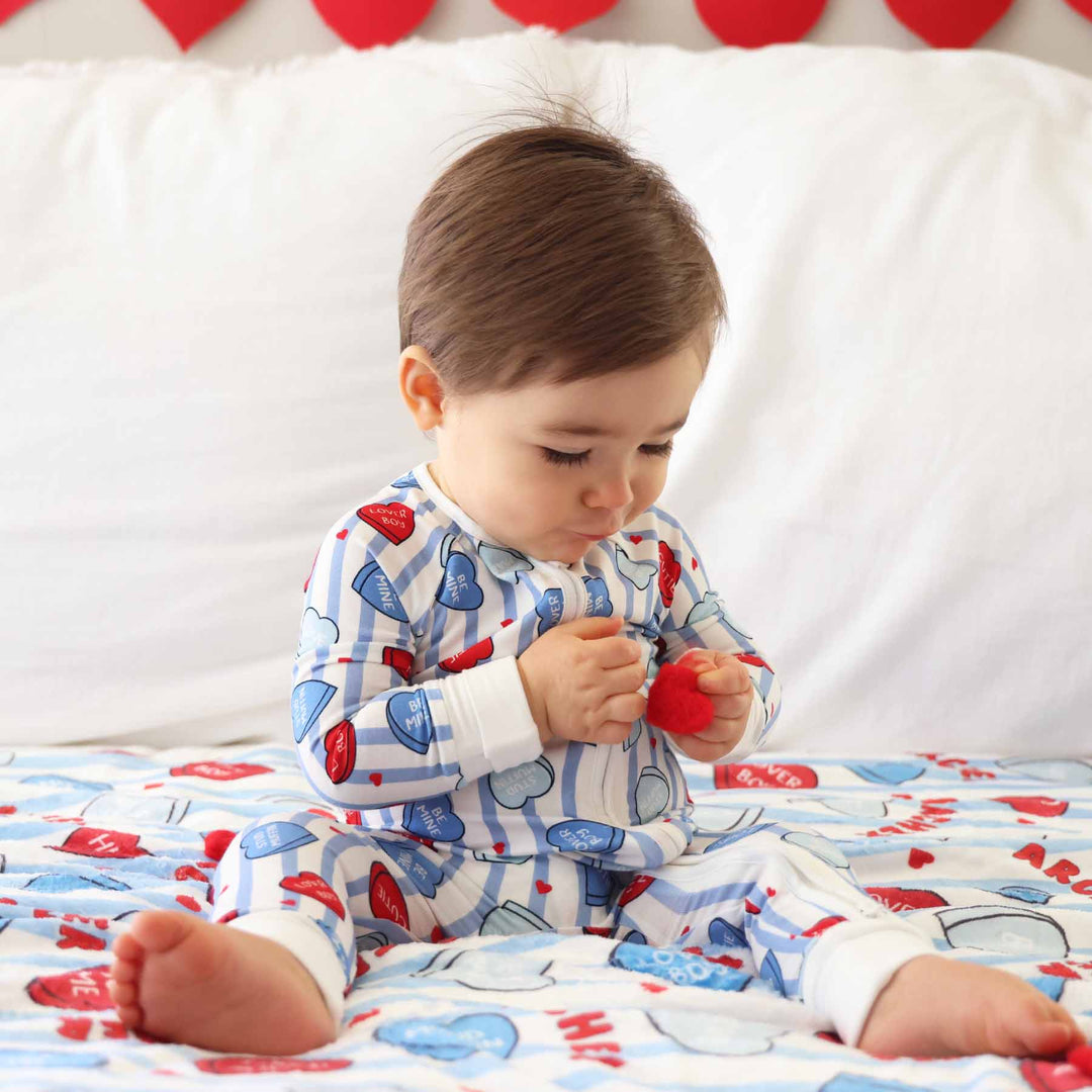 boy sitting on a bed in blue and white stripe candy conversation heart valentine's day pajama romper holding a tiny red heart 