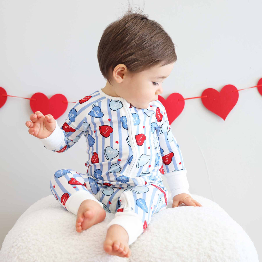 baby boy sitting on an ottoman wearing a blue striped candy heart pajama romper for valentine's day 