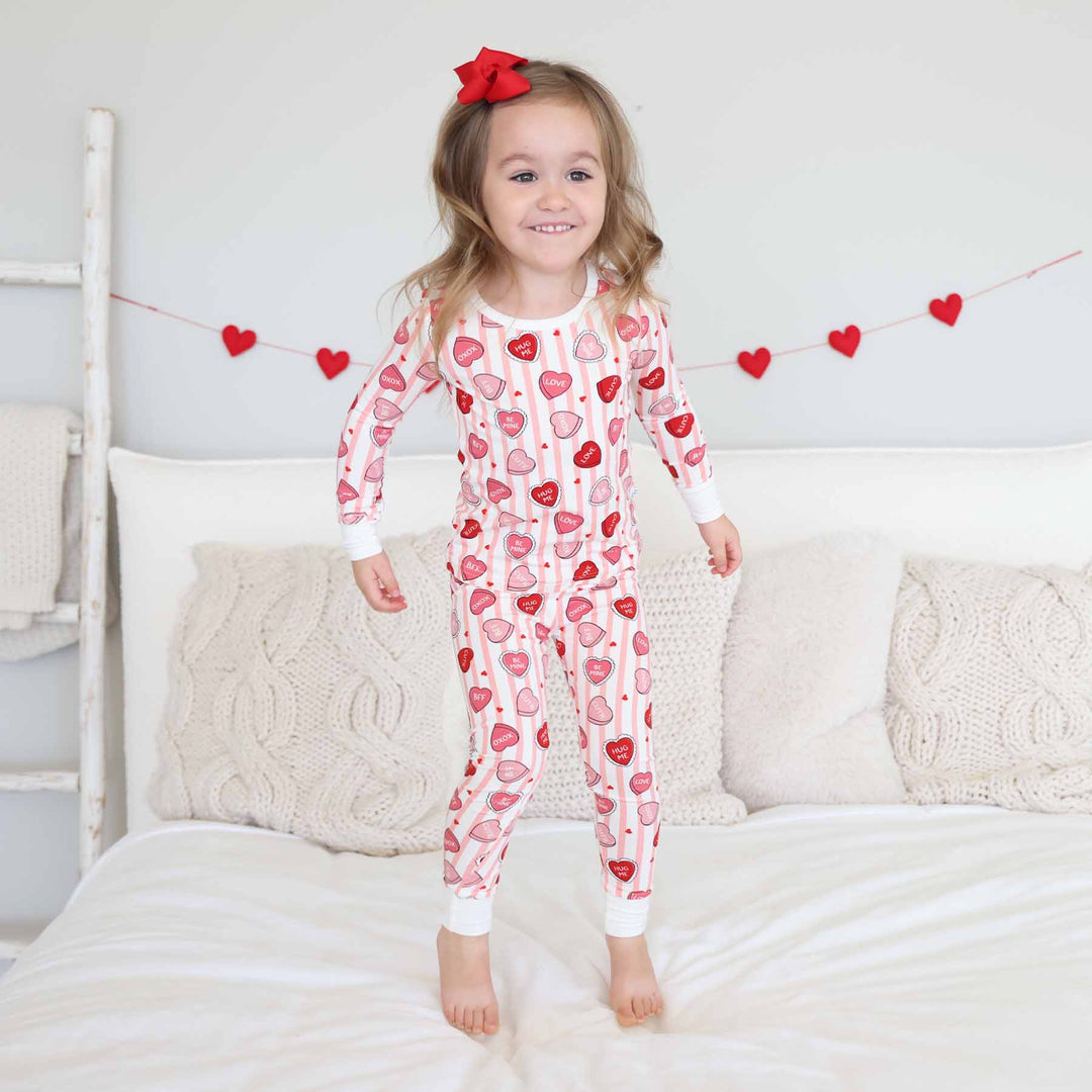 girl jumping on the bed in a pink stripe and candy heart pink and red conversation heart candy bamboo viscose two piece pajama set 