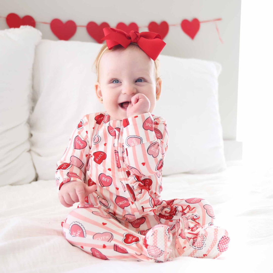 baby girl sitting on bed sucking her thumb wearing a pink stripe and candy heart valentine's day ruffle zipper footie 