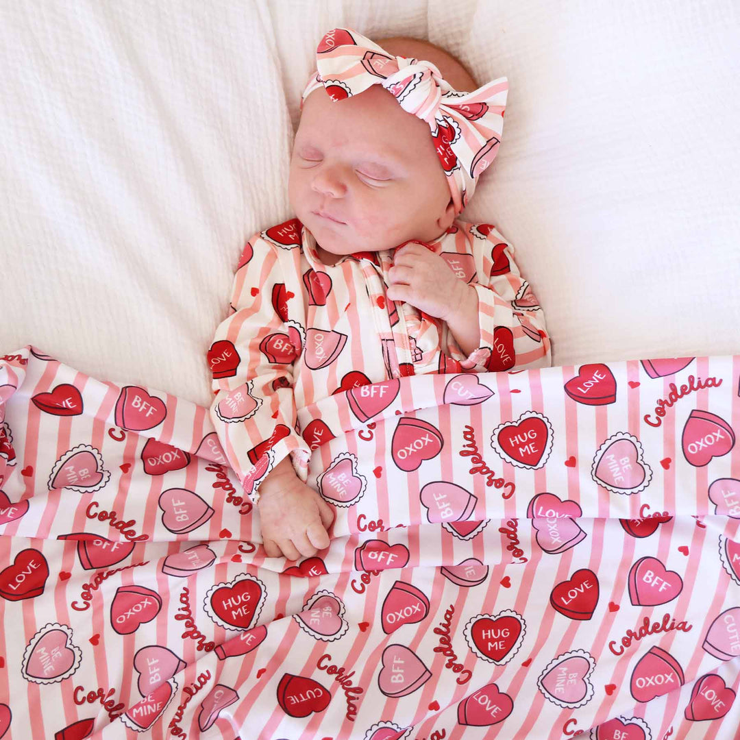 baby girl wearing the sweet like candy ruffle zipper footie with a matching headwrap with the personalized swaddle blanket on top of her 