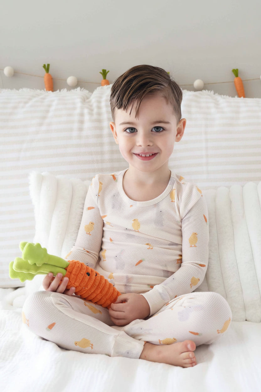 boy sitting on bed wearing a bamboo waffle neutral easter pajama set with chicks, carrots and bunnies holding a stuffed carrot 