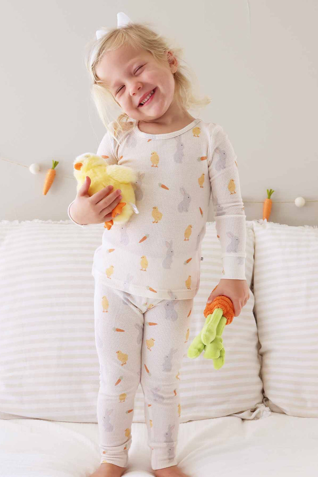 girl standing on bed wearing a neutral two piece pajama set with chicks, carrots and bunnies 