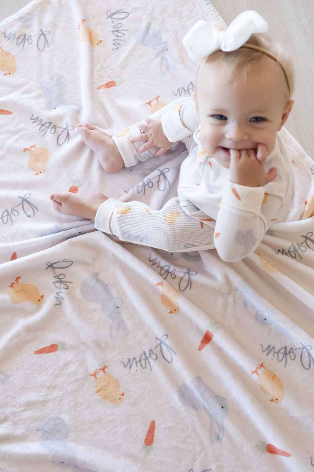 baby girl sitting on neutral easter personalized blanket wearing a matching zip romper pajama and white ribbon bow headband 