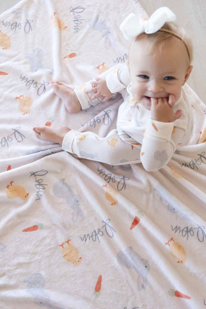baby girl sitting on neutral easter personalized blanket wearing a matching zip romper pajama and white ribbon bow headband 