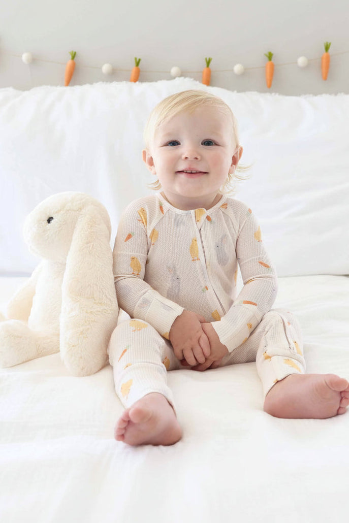 baby boy wearing sweet peep convertible zip romper pajama next to a white stuffed bunny animal with carrot garland in the background 
