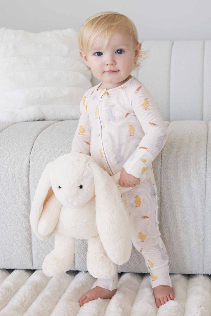 standing baby boy holding a stuffed bunny wearing a chick, peep and carrot bamboo waffle convertible zip romper pajama for easter 
