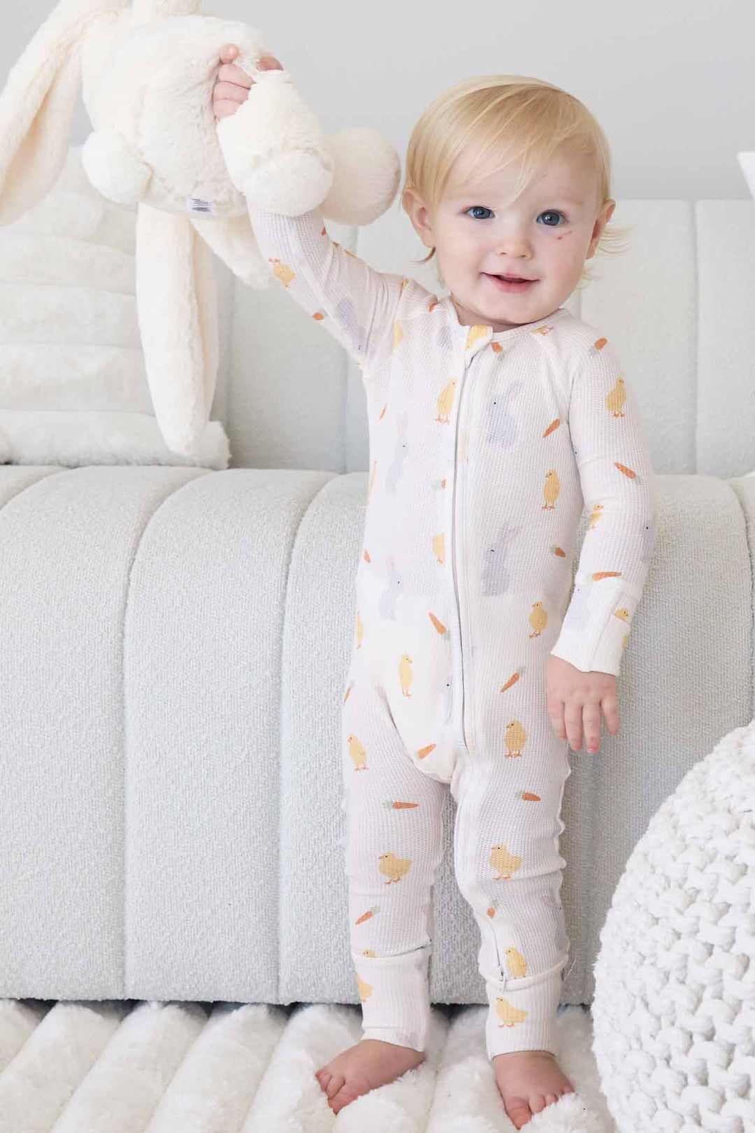 standing boy wearing neutral easter bamboo waffle zip romper pajama holding a stuffed bunny in the air 