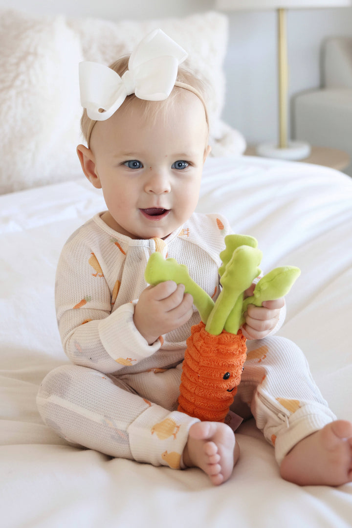 baby girl wearing the sweet peep bamboo waffle convertible zip romper pajama with a white ribbon bow headband and stuffed carrot 