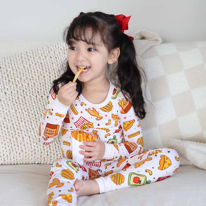 girl sitting on bed in fast food themed two piece pajama set for kids biting fake fry