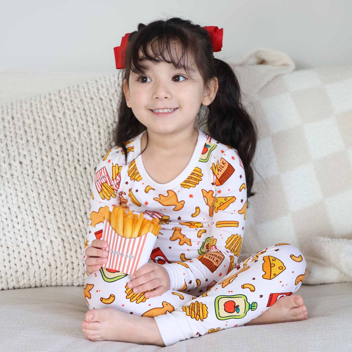 girl sitting on bed wearing a dino chicken nugget, mac and cheese and french fry pajama set holding caden lane french fries 