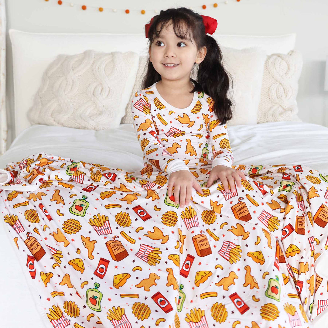 girl sitting on bed in a fast food themed two piece pajama set with a bamboo blanket draped over her lap 