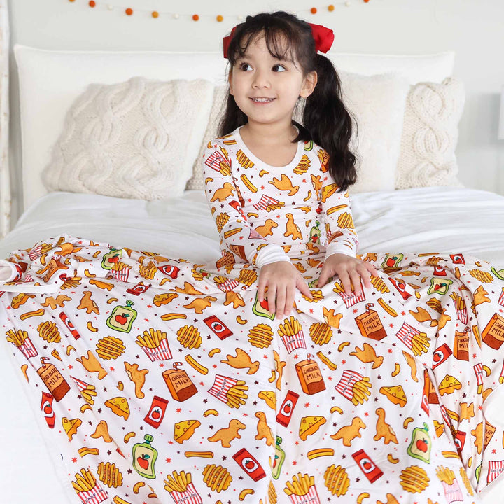 girl sitting on bed in a fast food themed two piece pajama set with a bamboo blanket draped over her lap 